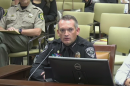 A police officer sits behind a microphone. The room behind is filled with chairs. 