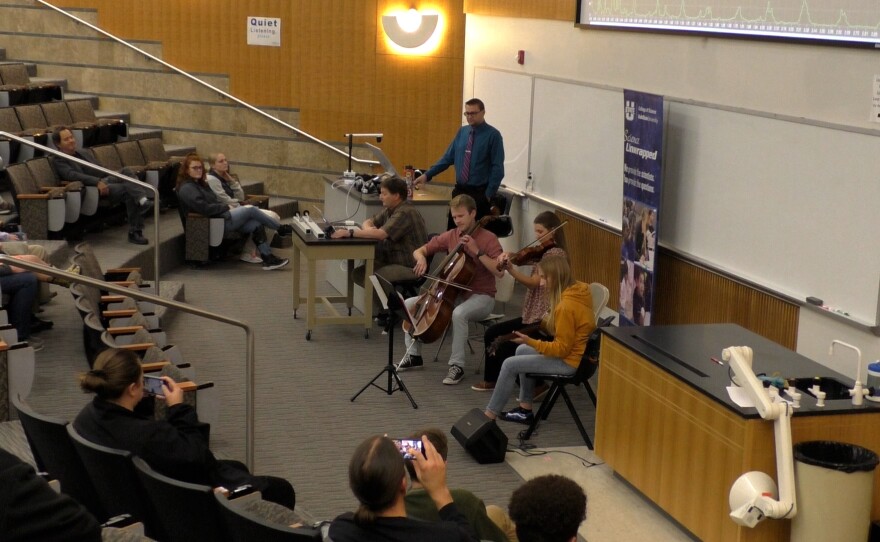 Three students play string instruments at the front of the lecture auditorium. The presenter stands to the side. Another man sits at a table between the presenter and the musicians with an oscilloscope to project a computer generated image of the music's soundwaves. 