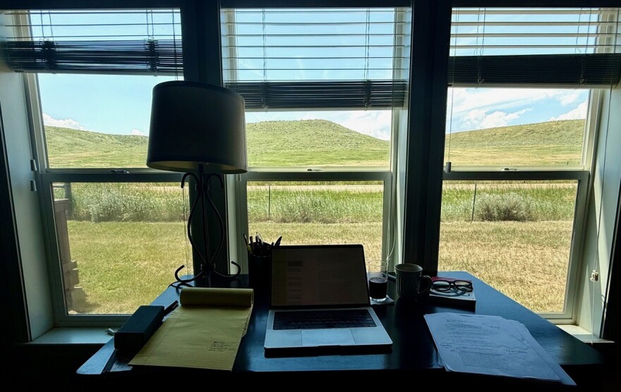 A desk with a lamp, notepad, laptop and sheets of paper, in front of three windows looking out onto a prairie with tall bluffs.