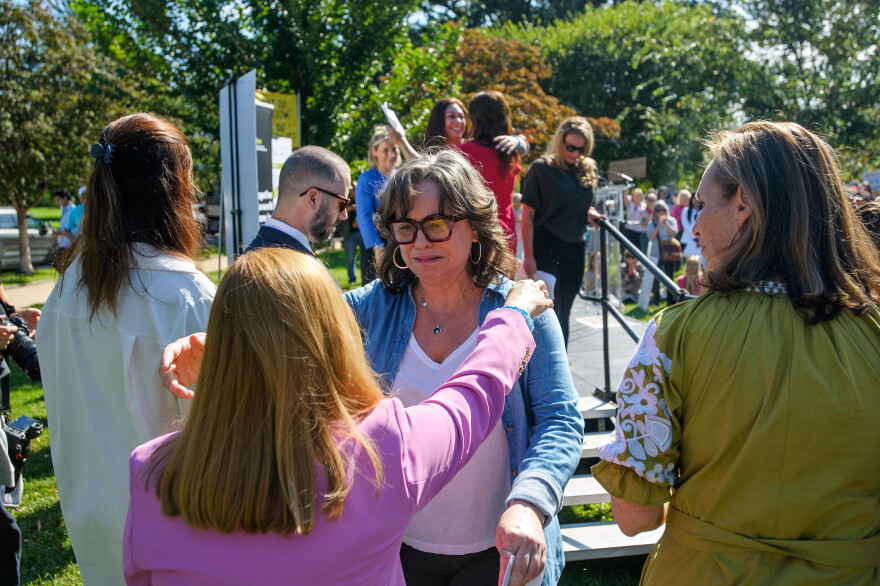 woman in blue jacket with white shirt underneath reaches arms out toward woman with pink or purple shirt and long red hair, who is turned away from the camera, on a sunny day, outdoors, with other people to their sides and in the background