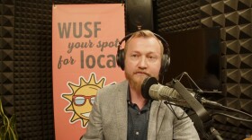 Man in suit jacket and button down shirt sits behind microphone with headphones on. A pink WUSF Your Spot for Local is behind him. 