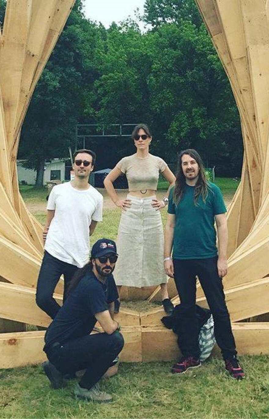 A woman and three men pose inside of an arched wooden art installation with trees in the background.