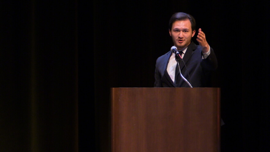 A young man in a suit extends his arm out to the crowd under a spotlight with a black background 
