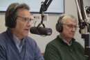 Two men sit in front of microphones with headphones one. One in a blue sweater is closer to the camera, the man next to him is in green. Each wear glasses.