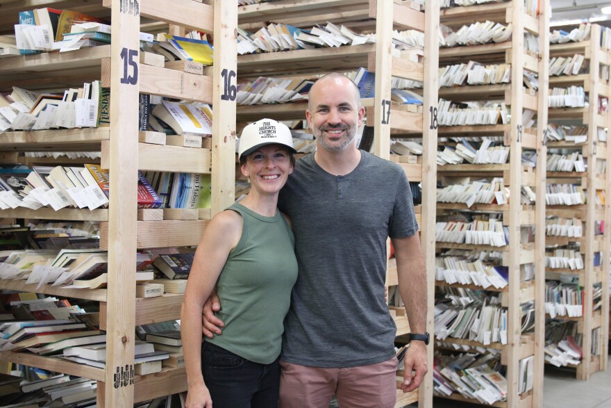 A woman in a green tank top and a white hat that reads "The Heights Church" smiles and side hugs a bald man with a grey beard, grey tee shirt and brown pants. Behind them are wooden bookshelves filled with books.