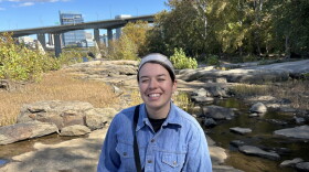 A young woman stands on a rock in front of a river. She is wearing a jean jacket, holding a water bottle, and smiling.