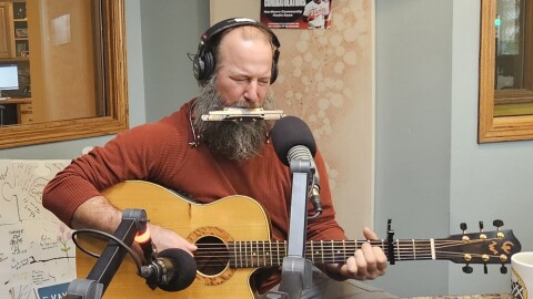 A man playing acoustic guitar and harmonica in a radio studio.