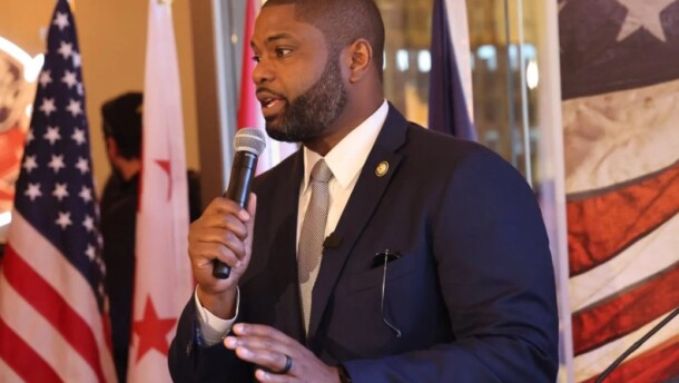 Black man with a beard and dark suit speaking into the microphone, looking to the left and an American flag to the left behind him