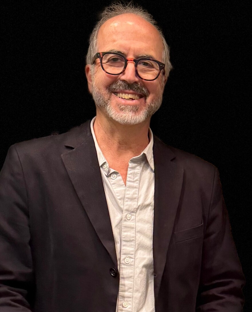 A man with a grey beard, wearing glasses, smiles at camera.