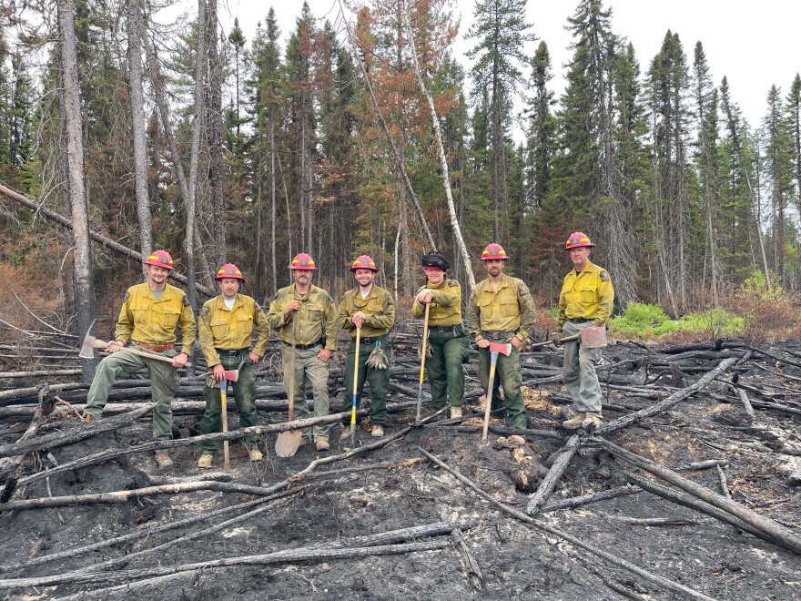  Seven men and women in green pants, yellow shirts, and orange helmets stand among burned forest debris in Quebec, Canada.  