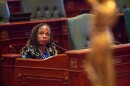 A woman with braided hair and a blue outfit sits at a wooden desk with a microphone in an Illinois lawmakers' assembly room, looking upward thoughtfully. Green chairs and ornate wood paneling surround her.