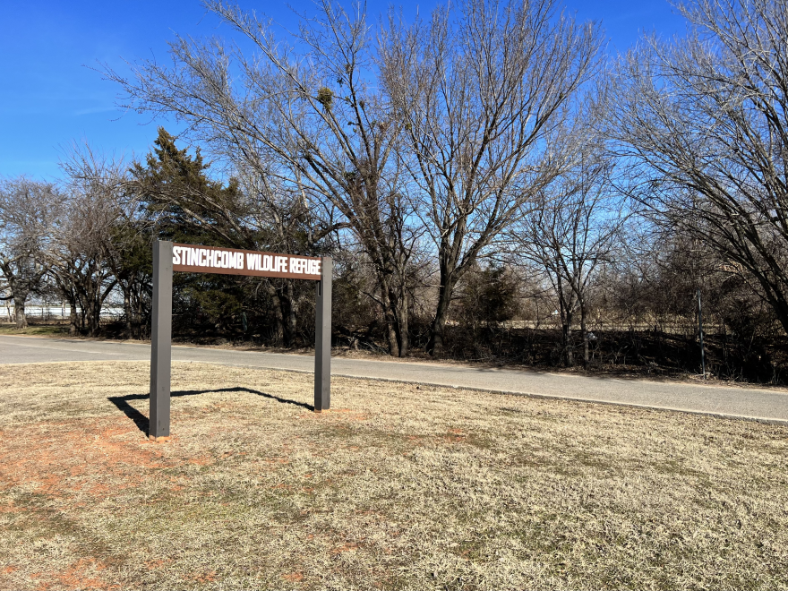 A brown sign with white lettering that says "STINCHCOMB WILDLIFE REFUGE." Behind the sign, a line of trees and blue sky. The trees are bare and the grass is yellow.