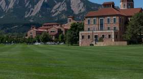 Tall brick buildings behind a large green field and the foothills of the Rocky Mountains.