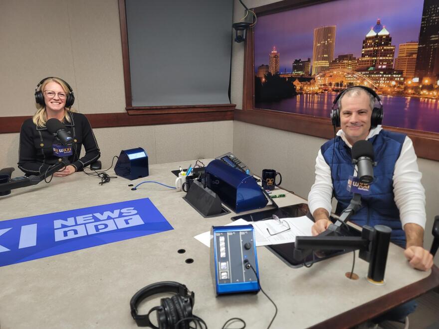 Two people wearing headphones sit at a table in a radio talk studio: a woman at left has long blonde hair and is wearing glasses and a long-sleeved black shirt; a man at right has short dark hair and is wearing a puffy navy vest over a white shirt.