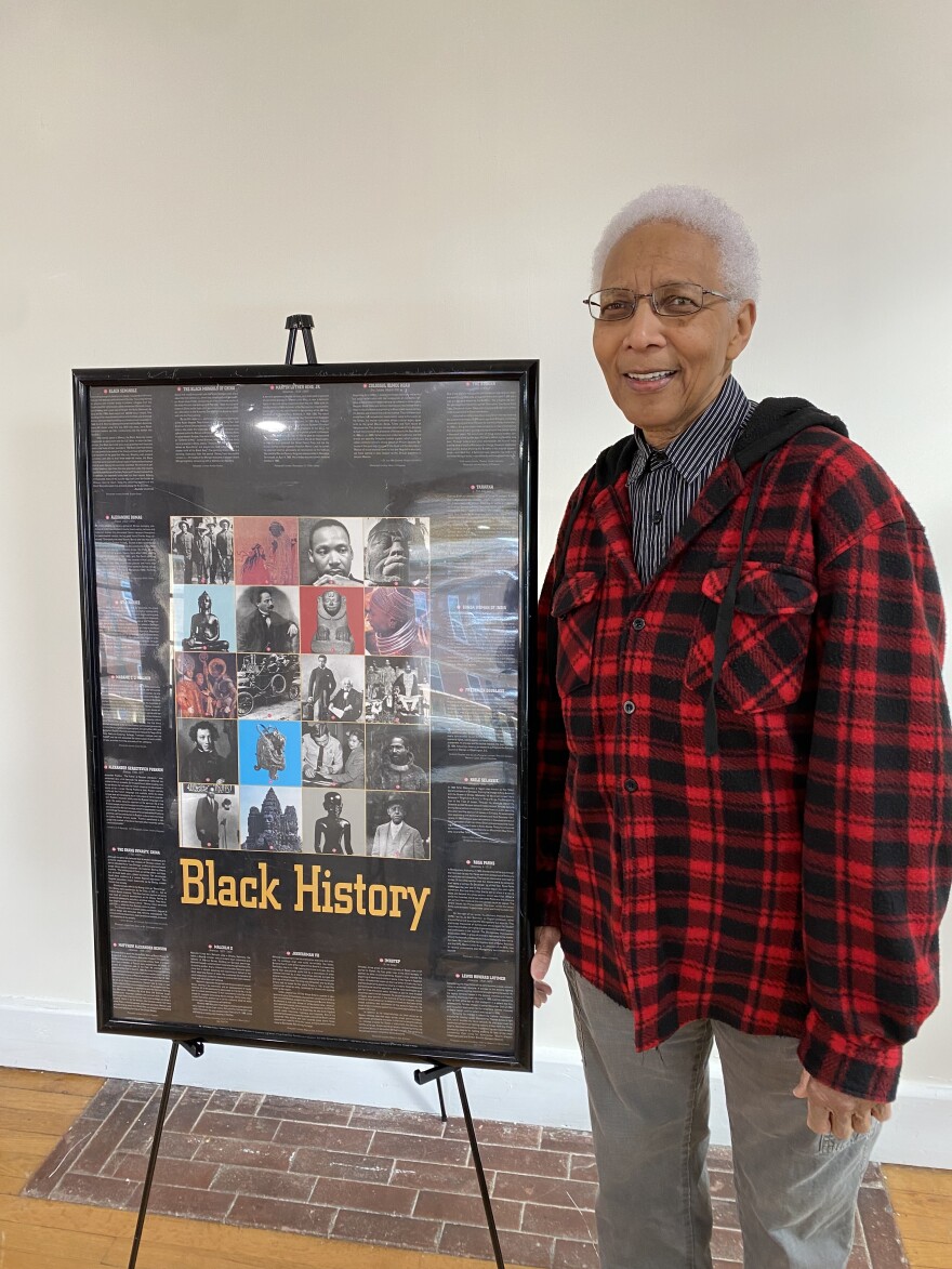 A woman with short grey hair stands in a red plaid shirt next to a Black History poster