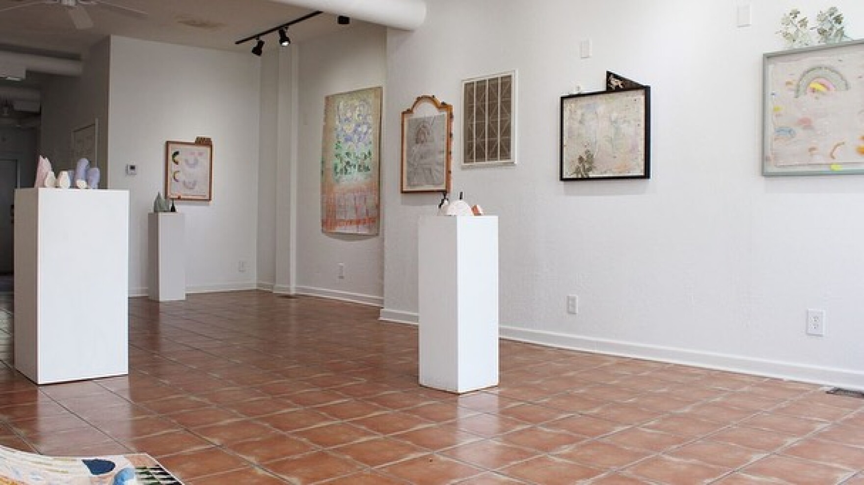 A white walled gallery space with brown tiled floors. Paintings are on the walls and there are three pedestals displaying small sculptures.