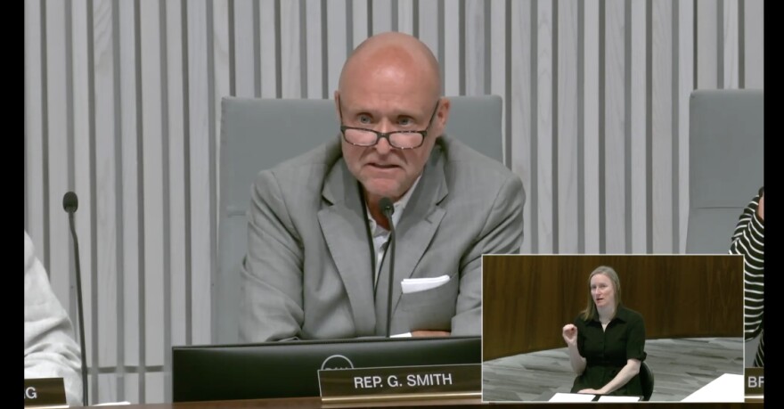 A middle aged bald man wearing a gray suit jacket and glasses sits at a dais and speaks into a microphone. 