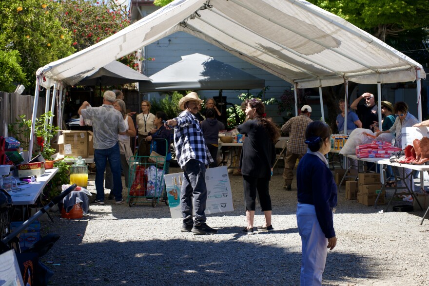 A man in a checkered flannel and straw cowboy hat points while talking to a woman dressed in a black long sleeve shirt and black cargo pants under a white tarp. Over ten people and many food, clothing, and home goods are scattered around them at a distribution event on a gravel backyard.