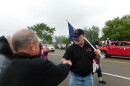A white man wearing a black polo shirt and baseball cap and carrying an American flag shakes the hand of another white man along a parade route.