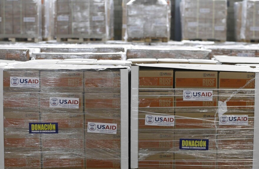 Humanitarian aid is stored in large boxes at a warehouse with labels that read 'USAID' and 'Donación 