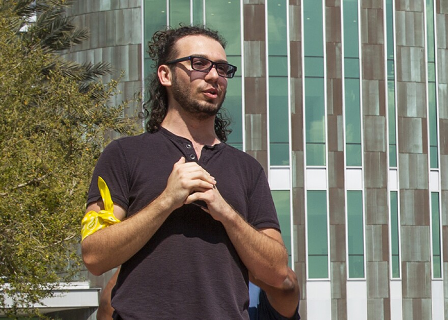 A curly haired man wearing glasses and a yellow bandana on his arm stands in front of the green student center. 