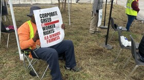 A person sits in a folding chair on yellowing grass, holding a sign that says "Corporate greed vs. worker rights: ILA demands fairness."