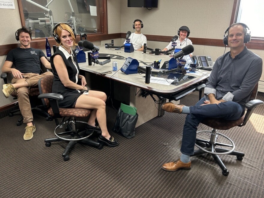 Five smiling people wearing headphones sit at a table in a radio talk studio: the man on the left in the foregound has short dark hair and is wearing a black t-shirt with khaki pants: the woman at the right in the foregound has red hair and is wearing a black sleeveless dress with white trim; the man on the left in the background has short dark hair and is wearing a white t-shirt; the person on the right in the background is wearing a white short sleeved button down shirt with a grey newsboy cap; the man at the end of the table has short dark hair and is wearing a grey long sleeved button down shirt with jeans and brown shoes. 