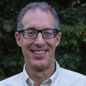 A man with gray hair and black glasses smiles at the camera, a blurry green background behind him.