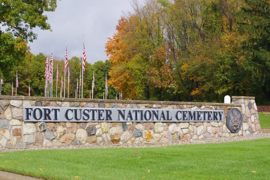 The entrance to Fort Custer National Cemetery, the sign and insignia on a stone wall with American flags along the entrance. The trees that line the road are in full autumn colors. 