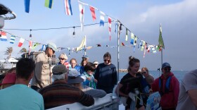 Flags strung around the top of the boat move in a slight breeze while many people stand and prepare to step off the boat. A woman holding a toddler under one arm and a dog under the other is stepping from the boat onto the dock while a man in a life vest holds open the boat's gate.
