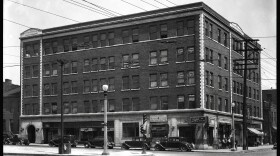 The Peoples Finance Building in Mill Creek Valley was among the buildings demolished in 1959. The black and white image is dated 1935, and photographed from a distance to show the brick, five-story building and five cars parked outside.  A group of people can be seen gathered on the sidewalk. 