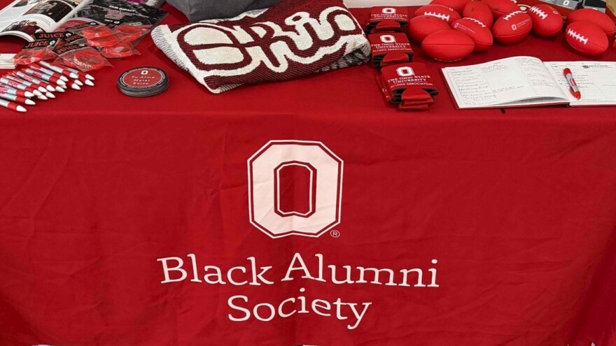 A close up of a table with a tablecloth that says "Black Alumni Society" under the Ohio State "O." On the table are pens, bracelets, koozies, and other items, all in Ohio State colors.