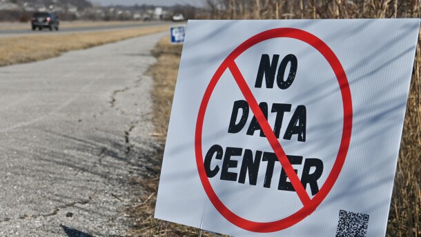 Exterior daytime image of a white sign stuck in the ground. It has a large red circle with a slash over the words "No Data Center." A highway can be seen in background where some cars are moving away.