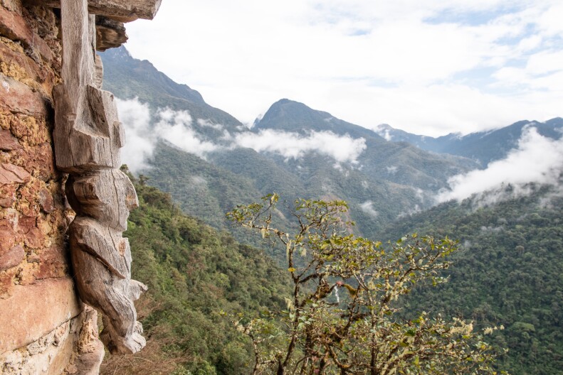 View of Rio Abiseo National Park: in the background mountain peaks covered in dense foliage in the mist. Left foreground, a wood carving of a human figure hanging from masonry or rack wall at an archaeological site. 