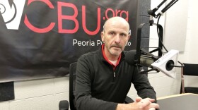 Peoria City Manager Patrick Urich sits at a table in front of a microphone in the WCBU master studio, with the station's brand logo on a banner in the background.  