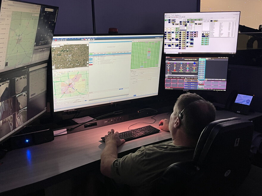 An emergency dispatcher sits in front of a bank of screens with maps and other information displayed.