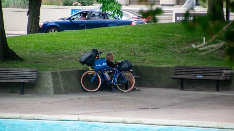 A man sits behind a bike with multiple bags strapped to it, avoiding the sun in Freimann Square.