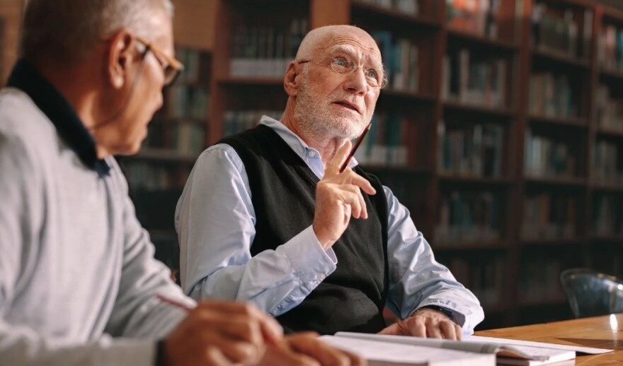Two older men sitting at a table with notebooks and bookshelves behind them