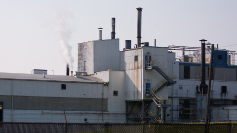 The Goodyear factory has a dark wire fence surrounding it, and is white and grey. The factory has a white puff of smoke coming out of the top of it. Rust runs down the side of some of the outer walls. 