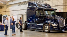People look at a dark blue semi truck with "Aurora" written on the door.