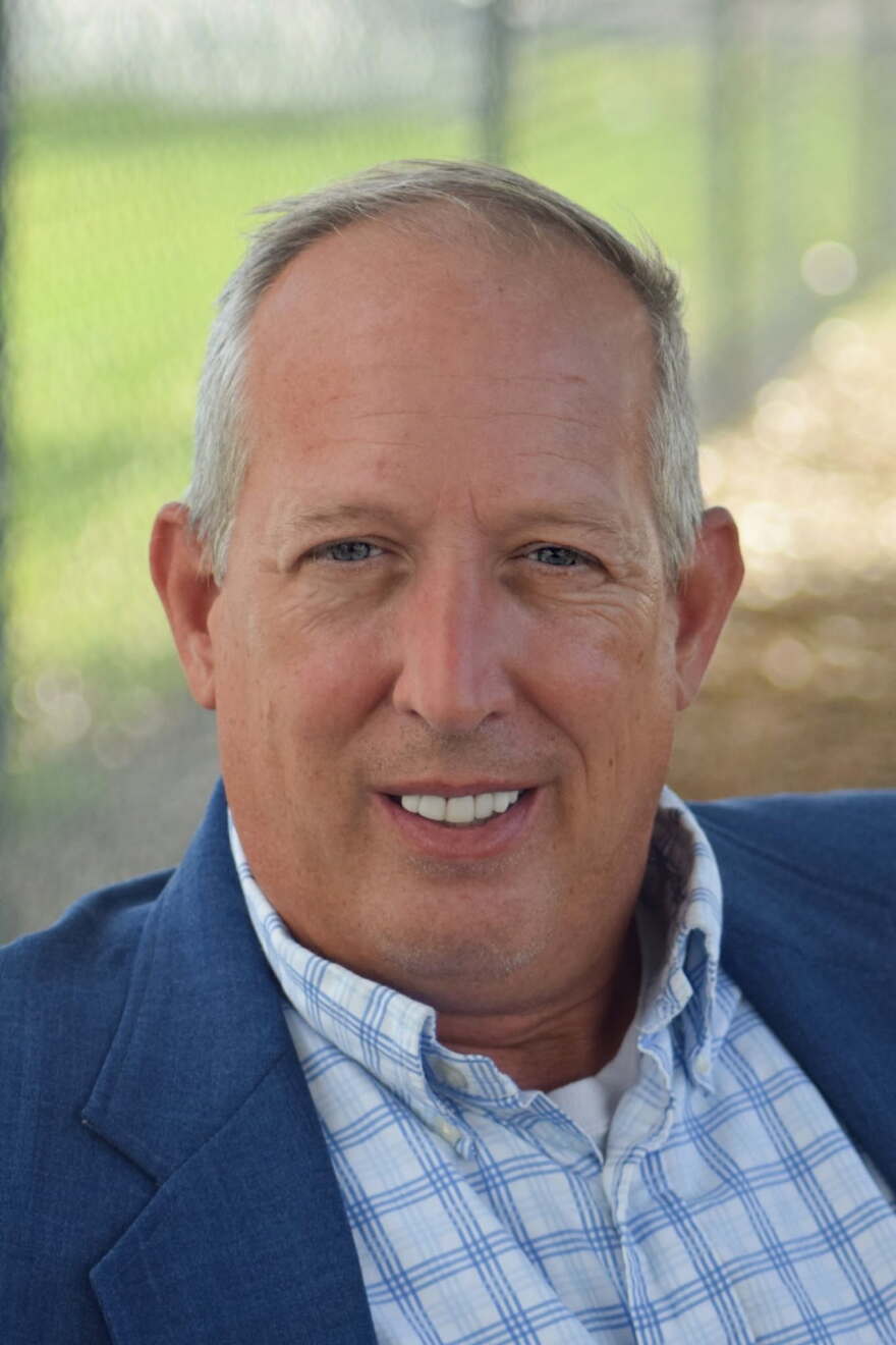 An older white man smiles in front of a fence.