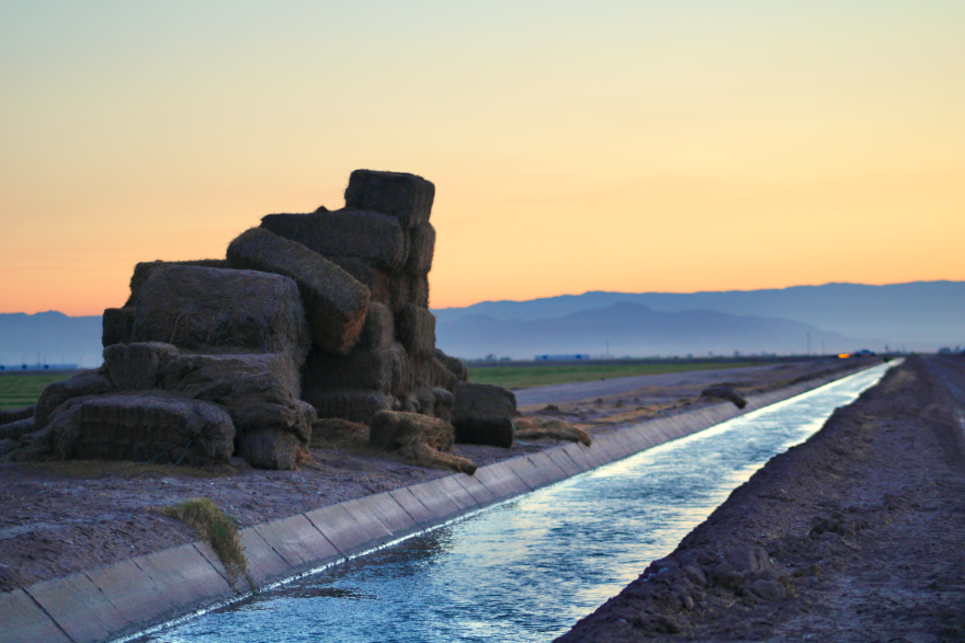 Stacks of hay bales sit near a narrow canal at sunset.
