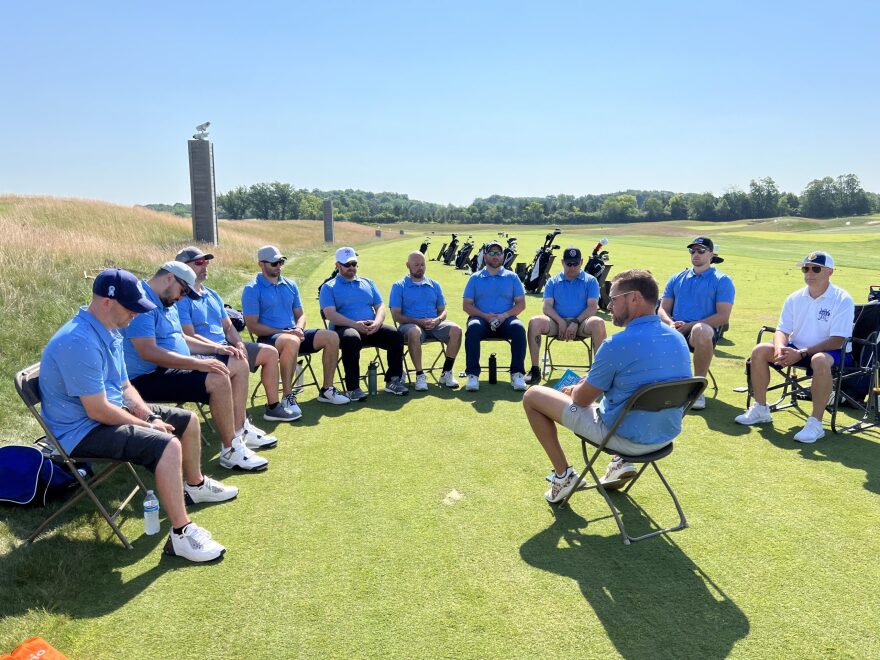 A group of men in blue shirts sit in a semicircle of chairs on a golf course around another man leading the conversation.