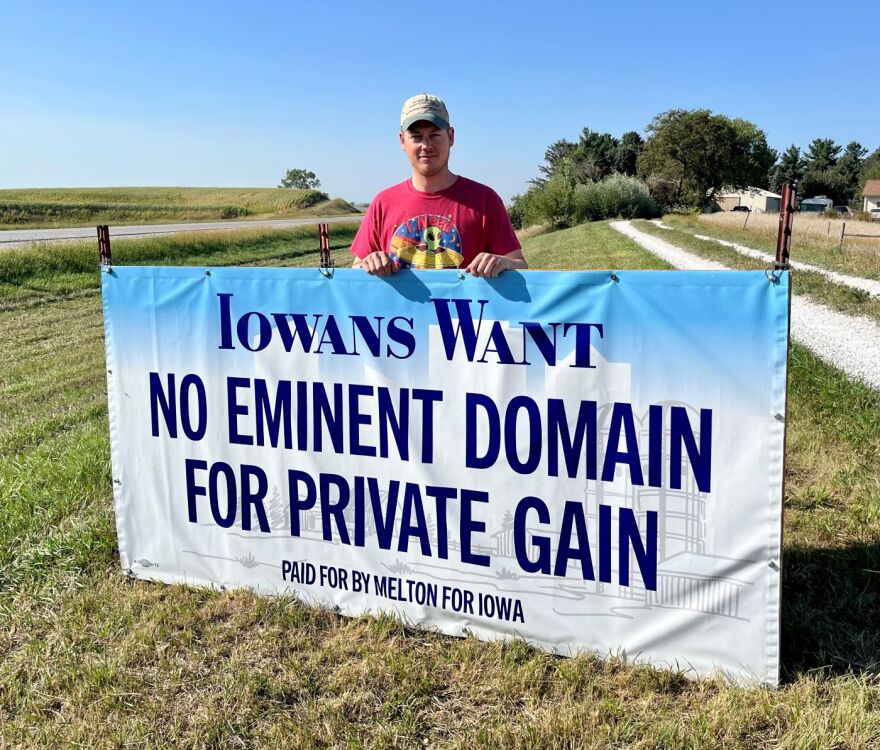 A man stands behind a sign that says "Iowans Want No Eminent Domain for Private Gain, Paid for Melton for Iowa. The man is standing next to a highway the sign is on grass with a gravel driveway to the right. He is wearing a red-ish shirt with an alien in a spaceship and a green-ish baseball cap.