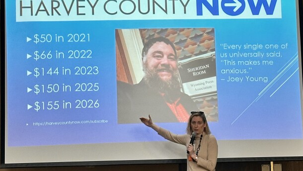A woman stands in front of a PowerPoint screen with her hand extended, speaking into a microphone. An image of a bearded man and some info about “Harvey County Now” is displayed behind her.
