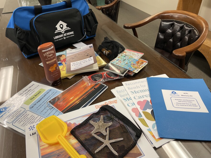 The ACPL beach-themed memory kit contains information for the caregiver, several books, photos with prompts on the back, colorful infographics, an empty sunscreen bottle, a puzzle, a plastic shovel, sunglasses and a few seashells.