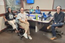 Five smiling people wearing headphones sit around a table in a radio talk studio: A woman front left has long black braids and is wearing white pants, black shoes, and a black graphic t-shirt with white text that reads "STREETS ARE FOR PEOPLE;" a woman front center has her dark hair tied back and is wearing sunglasses, a blue and white collared button-down shirt, tan pants, and white sneakers; a man back left is bald and is wearing a green graphic t-shirt; a woman back center has short black hair and is wearing a blue top; a man at right has very short dark hair and is wearing a black button-down shirt, jeans, and brown sneakers