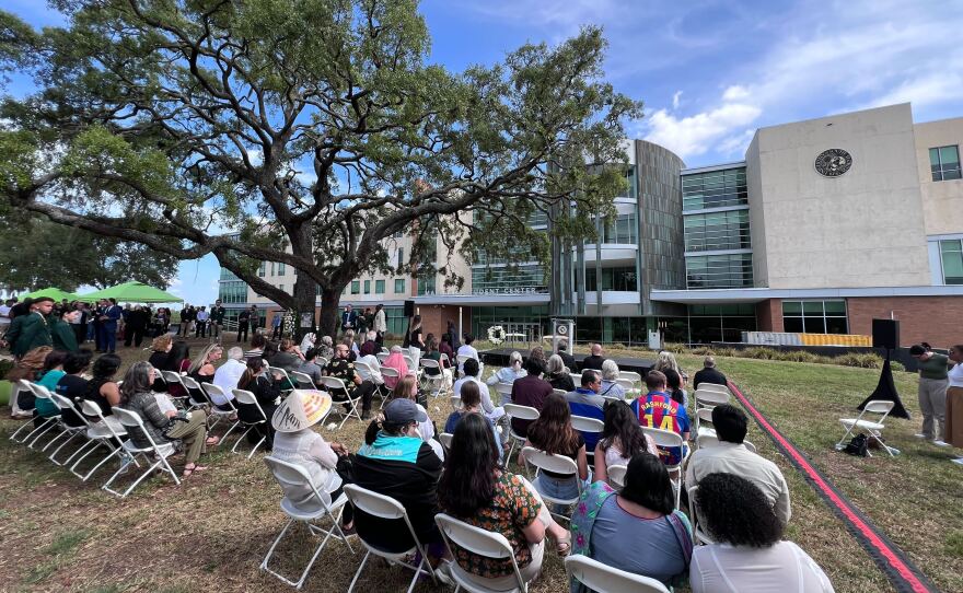 People sitting in rows of white chairs with people standing around them. They're facing a small, makeshift stage by a tree. 
