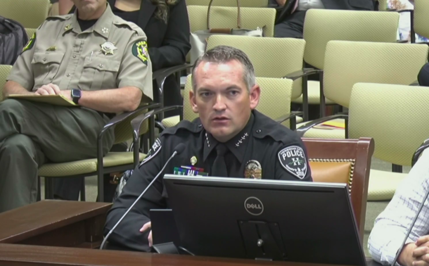 A police officer sits behind a microphone. The room behind is filled with chairs. 