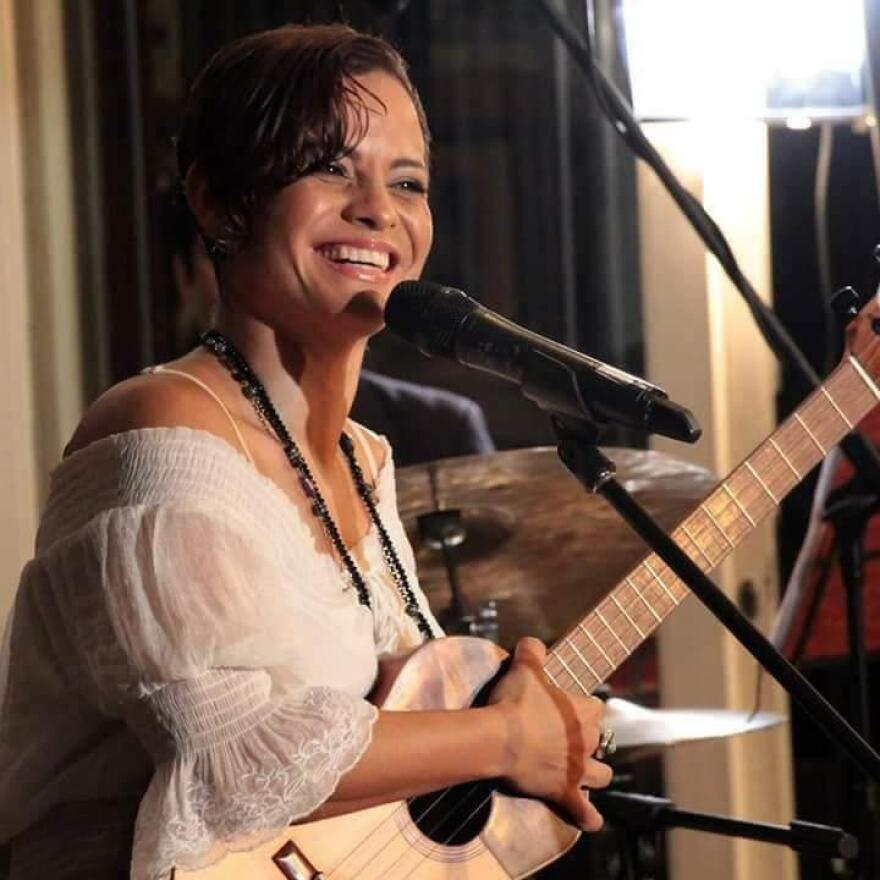 A woman smiles, singing into a microphone with a guitar in her lap.